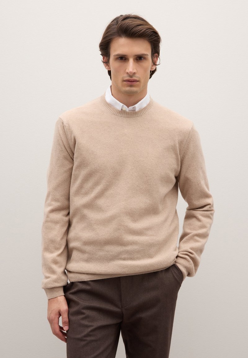Maglione beige in lana con scollo rotondo, polsini e orlo a coste, indossato sopra una camicia bianca con colletto, abbinato a pantaloni marrone scuro.