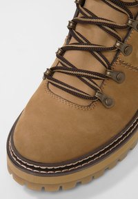 Hnědá kožená turistická bota s texturovaným povrchem, pevnými kovovými oky, hnědými tkaničkami a silnou, robustní gumovou podrážkou s prošívaným detailem.