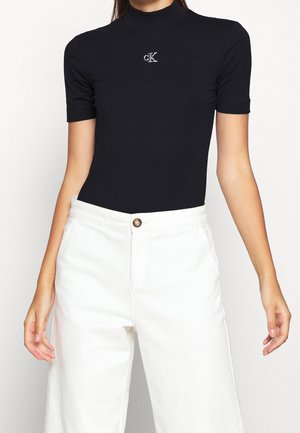 Vrouw draagt een zwarte coltrui met korte mouwen en zilveren "cK"-logo en witte, hoog getailleerde wijde broek met knoopsluiting.