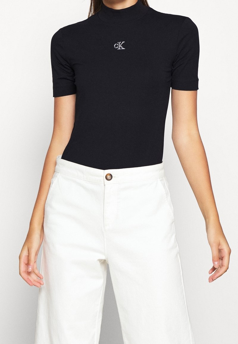 Kobieta ubrana w czarny, krótkorękawowy golf z srebrnym logo "cK" oraz białe szerokie spodnie z wysokim stanem, zapinane na guziki.