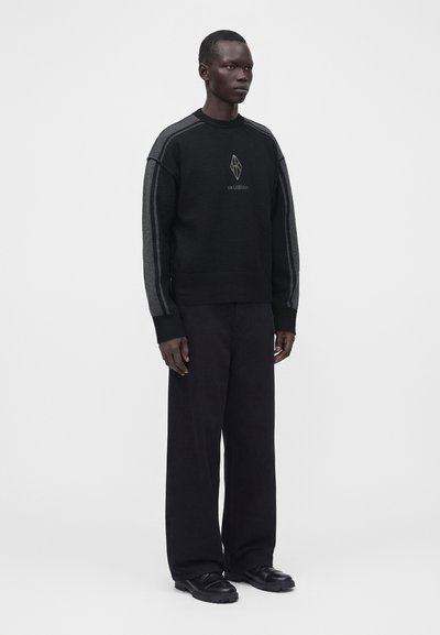 Homme portant un pull noir avec des rayures grises sur les manches, un pantalon noir à jambes larges et des chaussures noires, debout devant un fond blanc uni.