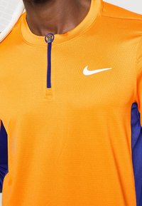 Nike træningsskjorte med orange krop og lilla sideskaller, med en kort zip-krave og et hvidt logo på brystet. Åndbart stof.