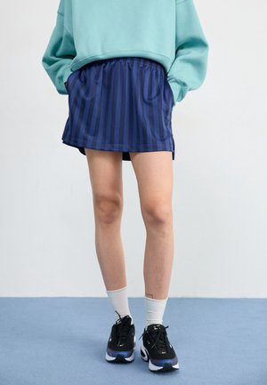 Personne portant une jupe rayée bleue, un sweat-shirt sarcelle avec les mains dans les poches, des chaussettes blanches et des baskets noires, debout sur un sol bleu.