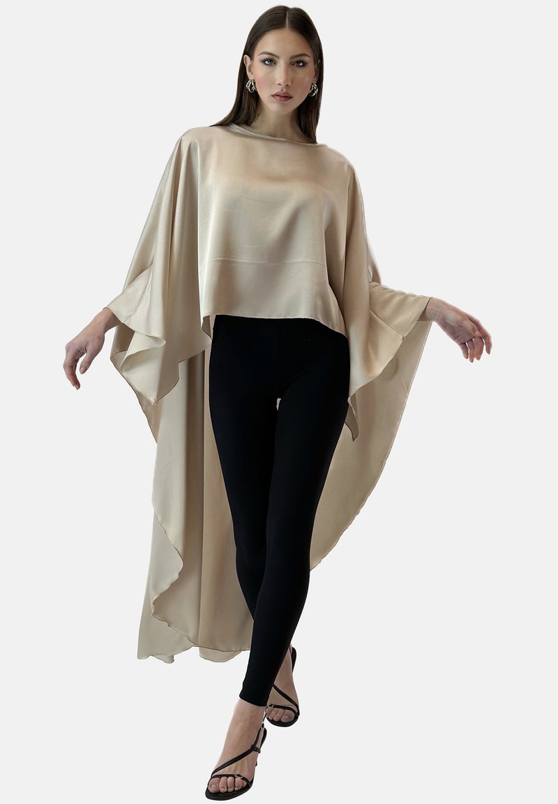 Beżowa satynowa bluzka z szerokimi rękawami, warstwowa konstrukcja, w połączeniu z czarnymi legginsami i czarnymi sandałami na ramiączkach, stojąca na tle jednolitym.