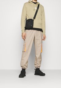 Felpa beige, t-shirt nera sotto, pantaloni cargo khaki con dettagli arancioni, borsa a tracolla nera e scarpe da ginnastica nere.