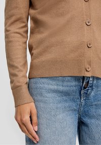 Brun stickad kofta med ribbade muddar och fåll, försedd med sex runda knappar. Kombinerad med ljusblå denimjeans. Mjuk textur.