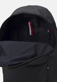Interno di uno zaino in tessuto nero con una texture liscia, dotato di un'etichetta con logo e di una tracolla con accenti rosso, bianco e blu all'interno. Chiusura con cerniera.