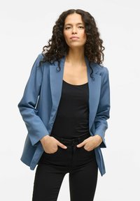 Modrý blazer se strukturovanými rameny, upraveným stylem, hladkým materiálem, otevřeným předním dílem a manžetami. Nositý přes černý top a černé kalhoty.