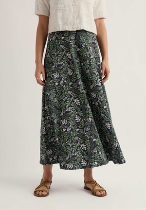 Frau trägt einen marineblauen Midirock mit Blumenmuster, eine beige kurzärmelige, geknöpfte Strickjacke und braune Sandalen mit Riemchen, steht vor einem schlichten Hintergrund.