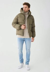 Olijfgroene gewatteerde jas met capuchon, gecombineerd met een kabelgebreide trui, lichtblauwe spijkerbroek en witte sneakers tegen een neutrale achtergrond.