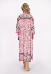 Kimono floreale lungo rosa con motivi intricati, bordi a contrasto e una cintura in vita. Tessuto morbido e maniche ampie. Abbinato a stivali beige.
