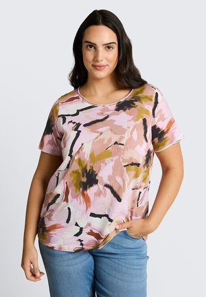 Kobieta z długimi ciemnymi włosami, ubrana w różową, czarną i oliwkową abstrakcyjną koszulkę z krótkim rękawem oraz niebieskie dżinsy, ręka w kieszeni.