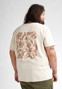 Beige T-shirt met een groot bruin bloemenontwerp op de rug, korte mouwen en een relaxte pasvorm, gemaakt van zacht katoen.