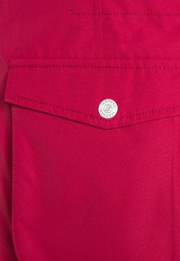 Czerwona tkanina o teksturowanej powierzchni, z kieszenią na klapę i metalowym guzikiem z logo. Wzdłuż krawędzi widoczne są przeszycia.