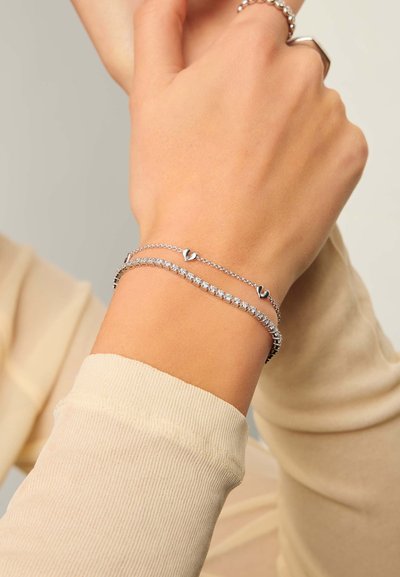 Χέρι με δύο ασημένια βραχιόλια, ένα με μικρά διακοσμητικά καρδιές και ένα με διαφανείς στρογγυλές πέτρες, πάνω σε φόντο μπεζ μπλούζας με μακριά μανίκια.