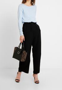 Woman wearing light blue long-sleeve top, black wide-leg pants, black high-heeled sandals, holding black handbag with snakeskin pattern sides.