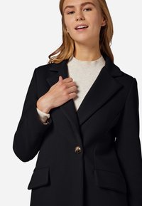 Schwarzer strukturierter, doppelt knöpfbarer Blazer mit Reverskragen, zwei frontalen Taschen und einem einzelnen goldenen Knopfdetail. Über einem hellen neutralen Pullover gestylt.