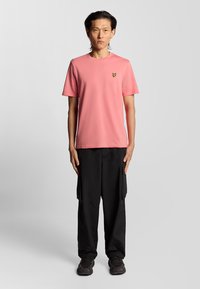 T-shirt rosa a maniche corte con un piccolo logo giallo sul petto, abbinato a pantaloni cargo neri e scarpe da ginnastica nere.