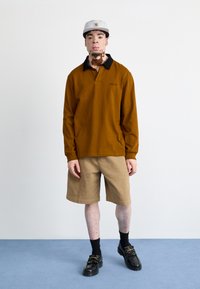 Long-sleeve brown polo with black collar, paired with khaki shorts. The model wears black shoes and a light gray cap, standing on a blue backdrop.