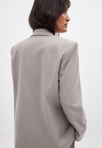 Gaišs pelēks žakets ar strukturētu dizainu, asi atlokiem un garām piedurknēm. Mīksta auduma, ar redzamām šuvju detaļām uz pleciem.