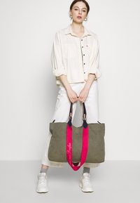 Grüne Canvas-Shopper-Tasche mit pinkfarbenen Trägern, verziert mit marineblauen Akzenten und gesticktem Text. Das Modell trägt ein weißes Hemd und helle Hose.