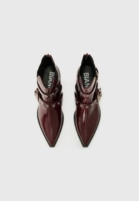 Bottines en faux cuir verni bourgogne, avec orteils pointus, présentant deux sangles décoratives avec des garnitures en métal argenté et des accents en métal rond.