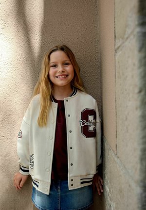 Uśmiechnięta dziewczyna o długich blond włosach nosi białą kurtkę varsity i jeansową spódnicę, opierając się o fakturowaną beżową ścianę.