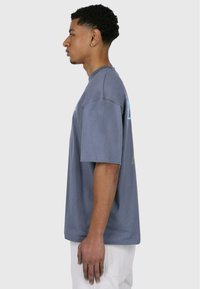 Grå bomulds t-shirt med korte ærmer, en afslappet pasform og sidesøm. Bemærkelsesværdig grafisk print i blå og grøn på ryggen.