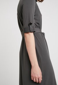 Šedé šaty s krátkým rukávem, obohacené o manžetu s kulatým akcentem. Měkká, hladká textura se štíhlým pasem a splývavým siluetou.