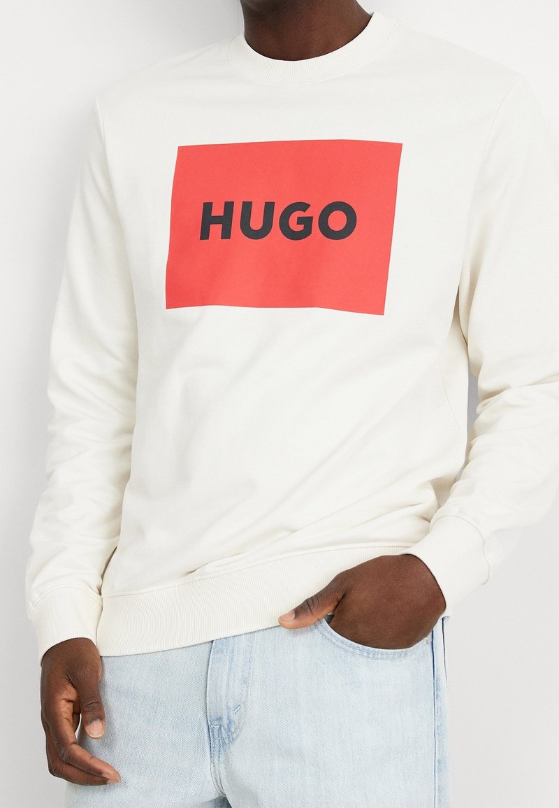 Felpa bianca a girocollo con una grande grafica rettangolare rossa che recita "HUGO" in lettere nere. Tessuto morbido, polsini e vita a coste.