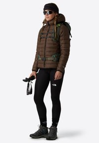 Brązowa kurtka puchowa z zielonymi akcentami, czarne legginsy i szare buty trekkingowe. Akcesoria to kije trekkingowe i okulary przeciwsłoneczne.