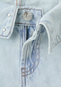 Jean en denim bleu clair avec fermeture éclair partiellement ouverte, bouton en métal gravé "AUTHENTIC DENIM MNG" et coutures orange visibles.