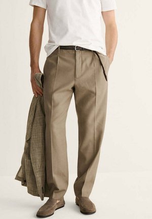 Hombre que lleva pantalones plisados beige, cinturón trenzado marrón, mocasines de ante marrones, camiseta blanca, sosteniendo un blazer marrón a cuadros en la mano izquierda.