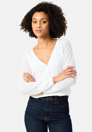 Witte sweater met lange mouwen en een diepe V-hals, met een effen lichaam en opengehaakte patroonmouwen. Gecombineerd met donkerdenim jeans.
