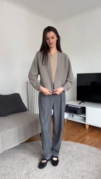 Kvinne justerer en grå cardigan over beige topp, har på seg grå bukser og svarte flate sko, står i en stue med sofa og TV.