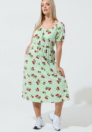 Femme en robe midi verte à pois blancs avec motif cerises rouges, posant les mains dans les poches, portant des baskets blanches sur fond blanc.