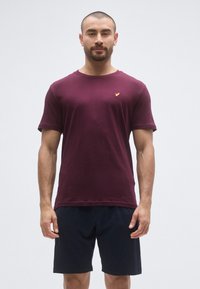 T-shirt col rond bordeaux en tissu doux, avec un petit logo doré sur la poitrine, assorti à un short bleu marine foncé.