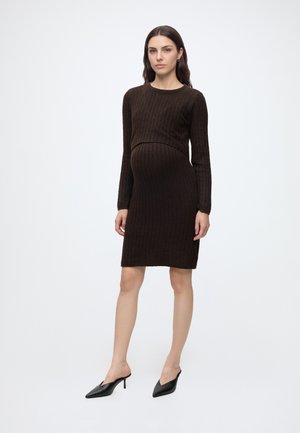 Femme enceinte portant une robe en maille marron foncé ajustée à manches longues et des mules à talons noires à bout pointu, debout sur fond blanc.