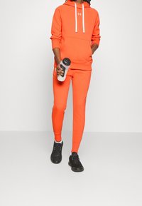 Orange hoodie med vita dragkedjor, matchande orange sweatpants och svarta träningsskor. Håller en svart och vit vattenflaska.