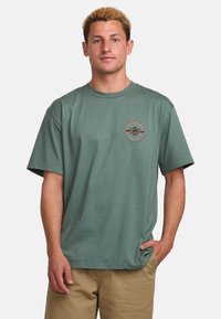 Grünes T-Shirt mit kurzen Ärmeln aus Baumwolle, featuring einen runden Halsausschnitt und ein kreisförmiges Logo mit Wellen auf der linken Brust.