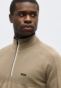 Νέος άνδρας κοιτάζει πλάγια φορώντας μπεζ ζακέτα με φερμουάρ σε ασπρόμαυρες ρίγες και λογότυπο "BOSS" στο στήθος.