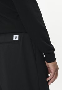 Čierne, ribované tričko s dlhým rukávom v kombinácii s úzkymi čiernymi nohavicami. Na zadnom vrecku je viditeľná etiketa s nápisom "M 73". Hladká textúra a priliehavý dizajn.