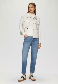 Weißer, strukturierter Weste mit zwei vorderen Taschen über einem hellen Oberteil, kombiniert mit blauen geraden Jeans und schwarz-cremefarbenen Sneakern.