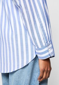 Bras et hanche d'une personne portant une chemise à manches longues rayée verticalement bleu et blanc et un jean en denim bleu clair.