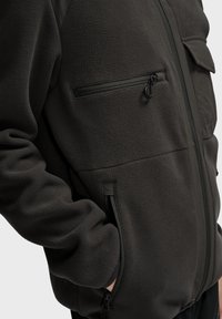 Mørkegrå fleecejakke med en lynlåslomme på brystet, sidelommer med lynlås og en struktureret overflade. Har et struktureret design og syede sømme.