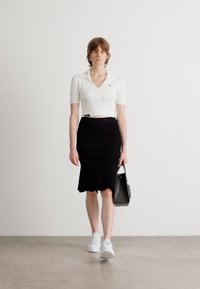 Weißes, enganliegendes Polo-Shirt mit kurzen Ärmeln, schwarzer knielanger Rock, weiße Sneaker und eine kleine schwarze Handtasche. Neutraler Hintergrund.
