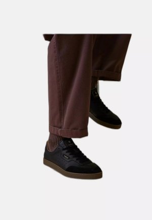 Person, die aufgerollte braune Hosen, braune gerippte Socken und schwarze Low-Top-Sneaker mit Gummisohlen vor weißem Hintergrund trägt.