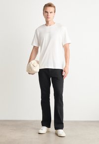 Hvit bomulls T-skjorte med brodert logo, svarte denimjeans og hvite sneakers, holder en beige myk veske med knapp.