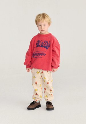 Jeune garçon blond portant un sweat-shirt rouge oversize et un pantalon beige avec des imprimés d'oiseaux et de champignons, debout sur un fond clair uni.