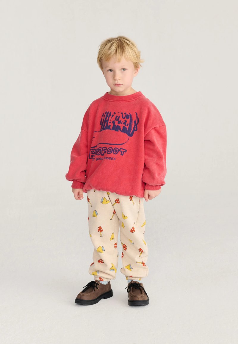 Jeune garçon blond portant un sweat-shirt rouge oversize et un pantalon beige avec des imprimés d'oiseaux et de champignons, debout sur un fond clair uni.